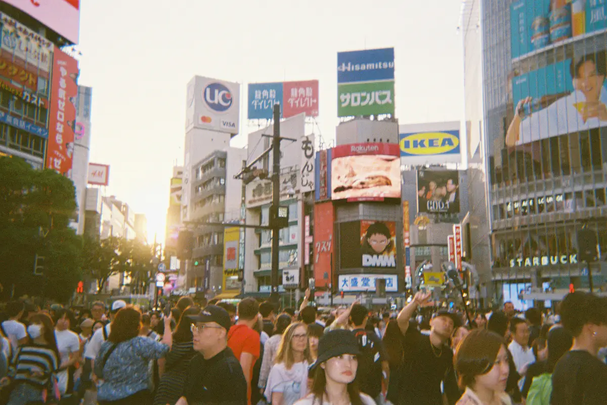 Pedestrians crossing Shibuya beneath bright screens at dusk.